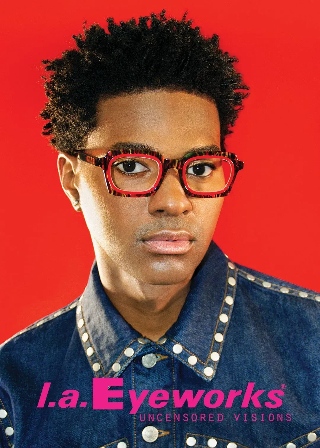 Person wearing red tortoiseshell glasses and studded denim shirt against red background