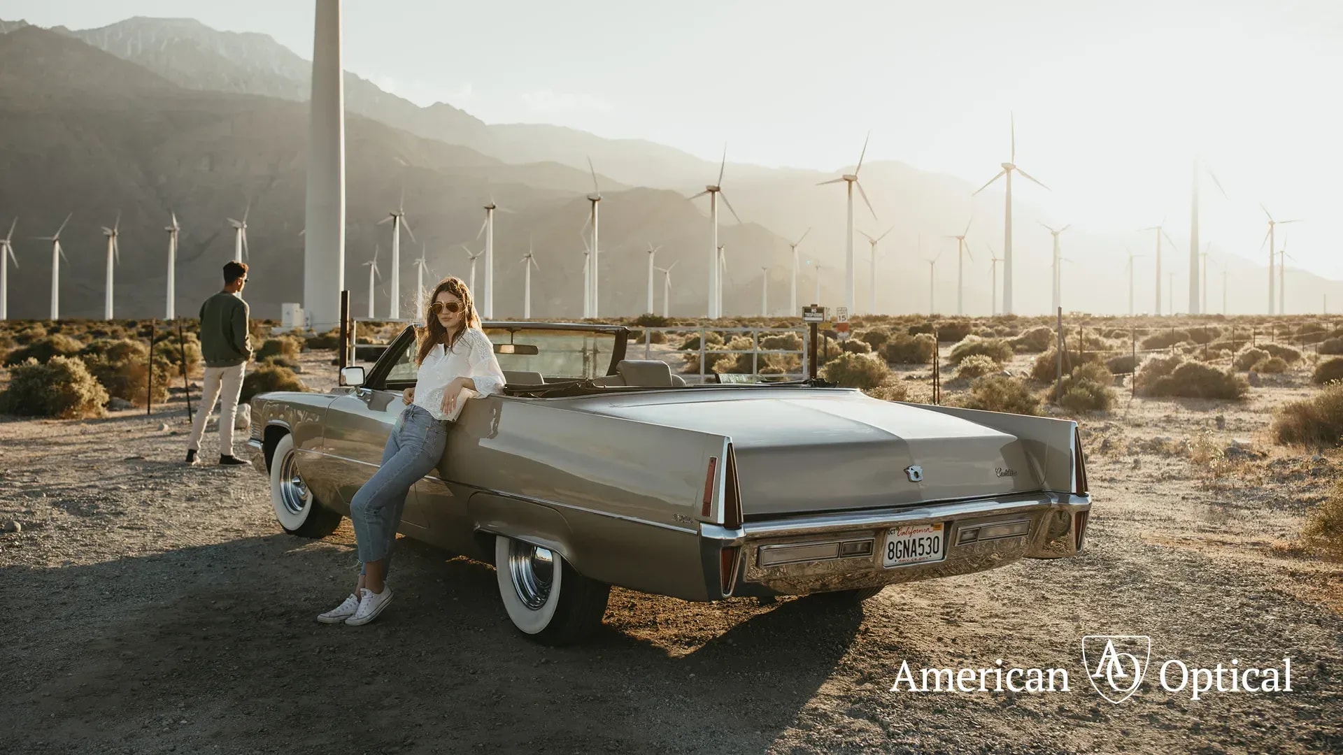 Woman in white shirt leans against tan convertible, surrounded by wind turbines under hazy desert sky